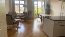 Open plan living and dining area with wooden floors, large windows, kitchen island with marble countertop, living room with armchairs, table with chairs, potted plants