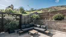Outdoor patio with wooden deck, black patio furniture including a sectional sofa and coffee table, surrounded by a wooden fence and lush greenery. In the background, there is a castle tower visible on a hill.