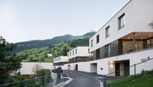 A modern, multi-story apartment building with balconies overlooking a scenic mountainous landscape. The building has a clean, minimalist design with large windows and a paved driveway leading to the entrance.