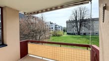 Balcony with a view of a grassy field and trees, red railing, beige shade