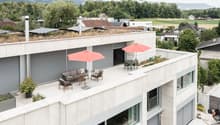 Modern white building with green roof terrace, solar panels, outdoor furniture, and an umbrella