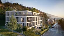 A modern, multi-story apartment building with balconies, surrounded by greenery and situated on a hillside overlooking a road and scenic mountain landscape.