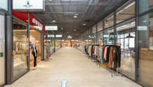 Shopping mall with glass walls, tiled floor, clothing stores, mannequins, clothing racks, and a blue arrow on the floor.