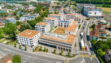 Aerial view of a modern office building complex with multiple buildings, green roofs, and solar panels, surrounded by trees and a road network.
