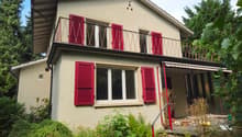 Two-story house with red shutters, beige walls, wooden terrace with a metal railing, awning over the entrance, and surrounded by greenery