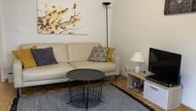 Beige sofa with patterned throw pillows, black and white coffee table, TV stand with TV, artwork on the wall, floor lamp, and a rug on the hardwood floor.
