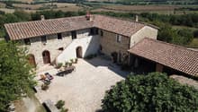 2-story stone farmhouse with red tile roof, courtyard with paved ground, outdoor seating area, surrounded by lush greenery and scenic countryside landscape