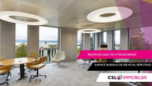 Large open office space with floor-to-ceiling windows offering a scenic view, modern furniture including round tables and swivel chairs, and colorful lighting fixtures on the ceiling.