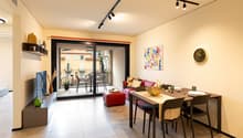 Modern open-plan living area, dining area with glass door leading to balcony, beige flooring, dining table, chairs, sofa, TV, painting, wall clock, ottoman, green vase, decorative items, view from balcony