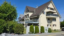 3-story residential building, beige exterior, brown roof, balconies on all floors, small garden, shrubs and trees in front