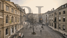 The image shows a historic city square with ornate buildings, cobblestone streets, and a lone tree in the center. There are people walking through the square, and the overall atmosphere appears to be one of a quaint, European-style urban setting.