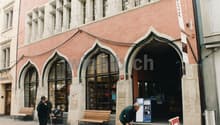 A building with arched windows and a large arched entrance, pink walls, wooden benches, and people walking by.