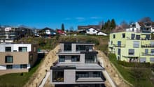 Modern house under construction, multiple levels, large windows, balconies, surrounded by other houses and greenery