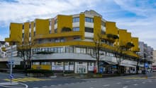 A yellow and white multi-story building with glass windows and balconies, located on a street with shops and trees.