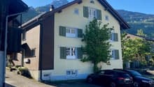 3-story residential building with a traditional Swiss architectural style, featuring a gabled roof, shuttered windows, and a balcony. There is a parking area in front of the building.
