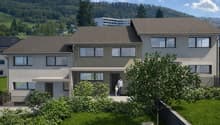 A contemporary two-story house with a brown roof, large windows, a small balcony, a spacious garden with a grassy lawn, surrounded by green foliage and a mountain range in the background.