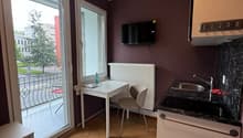 small room with a table, chair, and a television, a kitchen with a sink, stove, and a small refrigerator, a balcony with a view of the outside