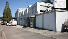White industrial building, flag on the roof, roll-up doors, garage door, vehicles parked in front, trees and mountains in the background