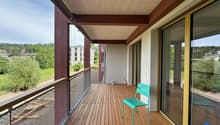 Wooden deck balcony with glass railing, overlooking a green, wooded area. The balcony has a turquoise chair and appears to be part of a larger residential building.
