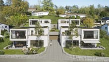 The image shows a modern residential complex with multiple two-story townhouse-style units. The buildings have a clean, minimalist design with large windows and balconies. The complex is surrounded by lush greenery, including trees and landscaped gardens.