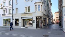 A three-story building with a shop on the ground floor, featuring a sign that reads 'RAHME-LADE'. The shop has large glass windows displaying various items. The building has a modern architectural design with white walls and green window shutters. There a