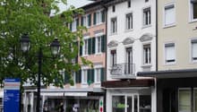 Three buildings with different colors, one with a balcony, shops on the ground floor, street lamp, tree, and bench