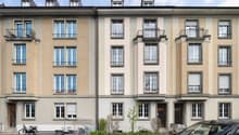 5-story building, beige and white exterior, multiple windows, balconies, front garden with plants and bicycles