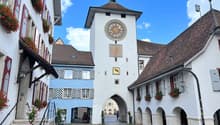 White building with clock tower, multiple windows with flower boxes, archway entrance, cobblestone street, archways along the building
