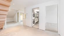 Unfinished attic space with exposed wooden beams, white walls, and a laundry room visible through the open doorway containing a washer and dryer.