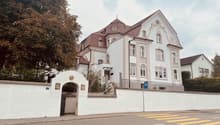 White house with brown roof, white walls, several windows, fenced off with white wall