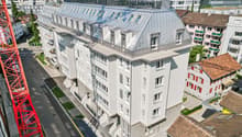 A modern white apartment building with a grey roof, multiple windows, and a metallic exterior. It is situated among other buildings in a city setting.