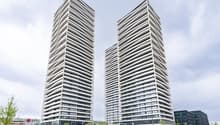 Skyscraper building, two tall buildings, concrete exterior, many windows, car park in front, street, trees, other buildings