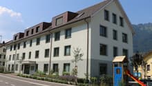 Apartment building with brown roof, many windows, front garden with plants and flowers, a child's playground, and mountains in the background.