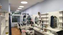 The image shows a room that appears to be a studio or office space. The room has gray walls, a large window, and various shelves and storage units along the walls. There are several mirrors and cosmetic items visible, suggesting it may be used as a beauty