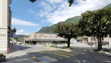 The image shows a street view of a town or city. There is a large supermarket building with the Migros logo visible. The street has a pedestrian crossing and parked cars. The surrounding area is lush with trees and greenery, and there are mountains visibl