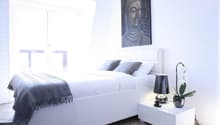 white bed, white nightstand, white lamp, white orchid, gray blanket, gray pillows, white walls, white ceiling, white floor, painting of Buddha