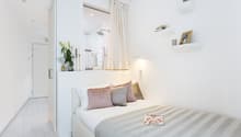 Modern small bedroom with tiled floor, a double bed with a gray and white striped bedspread, a nightstand with magazines, built-in wardrobe, a curtain for the shower, shelves with decorative items, a plant, and a decorative lamp.