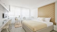 Modern bedroom with beige bed, white headboard, two white armchairs, round glass table, decorative rug, large windows with white curtains, and a desk with a television on the wall.
