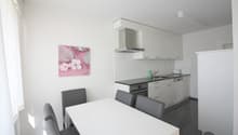 white tiled walls, kitchen with black countertop, white cabinets, modern appliances, grey chairs, dining table, artwork on wall, window with white curtains