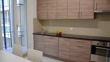 Modern kitchen with wood-paneled cabinets, black countertop, stainless steel sink, and a dishwasher. Large windows provide natural light and a view of the exterior. The kitchen also includes a dining area with white chairs.