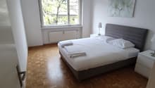 Spacious bedroom with large window overlooking trees, hardwood parquet flooring, grey upholstered bed frame, and a decorative floral painting on the wall.