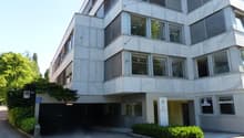 A multi-story office building with a garage entrance, large windows, and a modern concrete and glass facade. The building is surrounded by trees and greenery.