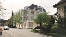 4-story apartment building with a modern design, gray exterior walls, and large windows. The building is surrounded by trees and landscaping, and there is a parking area in front of the building.