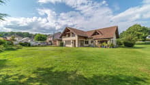 Detached house, brown roof, beige walls, windows with wooden frames, car in the driveway, large green lawn, several bushes and trees, cloudy sky