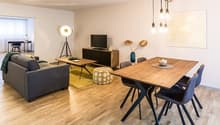 Open-concept living and dining area with hardwood floors, gray sofa, wooden dining table and chairs, TV stand, and decorative wall art