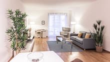 large windows, large gray couch, coffee table, glass table, armchair, desk, chair, two potted plants, wooden floor