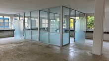 The image shows a large, open office space with concrete floors and glass partition walls. The space appears to be in a state of renovation or construction, with some debris on the floor. The glass partitions divide the space into separate rooms or areas,
