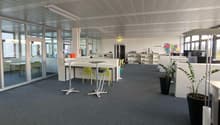 The image shows a modern and open office space with a high ceiling, glass walls, and a mix of workstations and lounge areas. The space has a gray carpet floor, white walls, and a gray ceiling with exposed beams. There are several desks with chairs, shelve