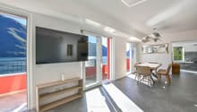 modern living room, gray floor, white walls, large window, flat screen TV mounted on wall, wooden table and chairs, dining area with open balcony, view of mountains and lake, decorative lighting