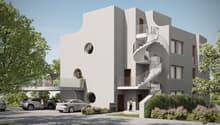 Modern, multi-story apartment building with white exterior, circular windows, and spiral staircases leading to balconies. There is a parking area in front of the building with several cars parked.
